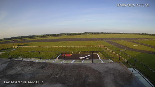 An outdoor view from a webcam at Leicestershire Aero Club showing the top of a helicopter’s tail rotor and tail fin in the foreground. Beyond, there is a grassy area with a small red and gray landing pad, a windsock on a pole, and a large open runway extending into the distance under a clear sky. The timestamp on the image is February 6, 2025, at 8:00 PM.