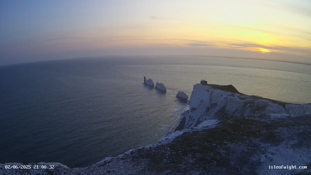 A view of a coastal landscape at sunset, with white chalk cliffs extending into the sea. There are several large rocks or small islands in the water near the cliffs. The sky is partly cloudy with the sun low on the horizon, casting a warm glow over the scene. A small building can be seen on top of one of the cliffs. The timestamp and website URL are visible in the bottom corners.