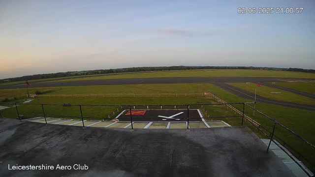 A view from a webcam shows an airfield with a large clock painted on the ground in the foreground, displaying the time as approximately 7:05. Beyond the clock area, there is a grassy field enclosed by a low fence, and a small red flag on a pole near the left side of the image. There are runways extending into the distance under a clear sky with a few clouds, and the horizon features some trees and fields. The timestamp on the image indicates it was taken on February 6, 2025, at 21:00:57.