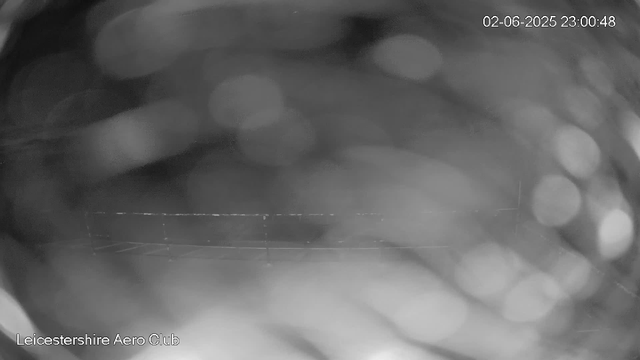 A black-and-white image captured by a webcam shows an indistinct, blurry scene with circular light spots scattered throughout. The background appears to be an indoor setting, with some faint outlines of objects and structures visible. The timestamp on the top right reads "02-06-2025 23:00:48," and "Leicestershire Aero Club" is written at the bottom left.