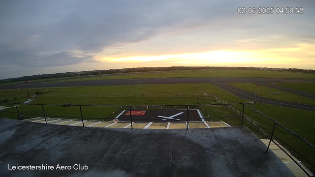 A view from a webcam at Leicestershire Aero Club showing an airfield during sunset or sunrise. The foreground features a helipad with a large letter "X" on it, surrounded by a safety railing. Beyond the helipad, the airfield has grassy areas, a runway, and some small structures or equipment. The sky is partly cloudy with a glow of orange near the horizon. The timestamp in the upper right corner reads "03-06-2025 04:59:52".