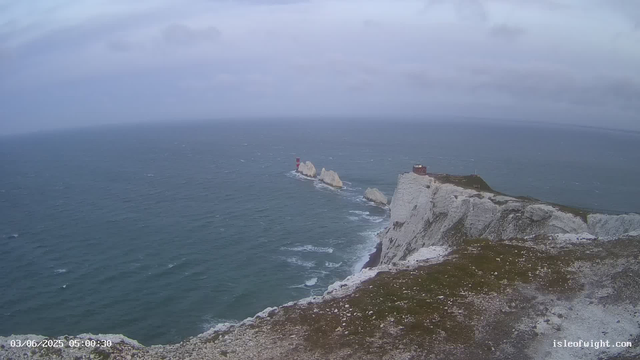An image of a coastal landscape shows white chalk cliffs rising from the ocean, with a small building or lighthouse on top of the cliffs. In the distance, there are two smaller rocky islands with a lighthouse on one of them. The sky is cloudy and overcast, and the ocean appears choppy with waves. The scene is from a webcam, timestamped "03/06/2025 05:00:30" and has a credit to isleofwight.com.