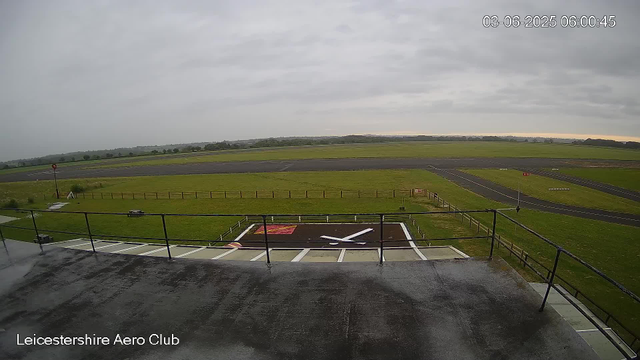 A view from a webcam at Leicestershire Aero Club showing the runways and grassy fields on a cloudy day. The timestamp in the upper right corner indicates the date and time as June 3, 2025, at 6:00 AM. The scene includes a helipad with a large white "X" marking, surrounded by a safety fence, and a small platform or balcony with a wet surface in the foreground.