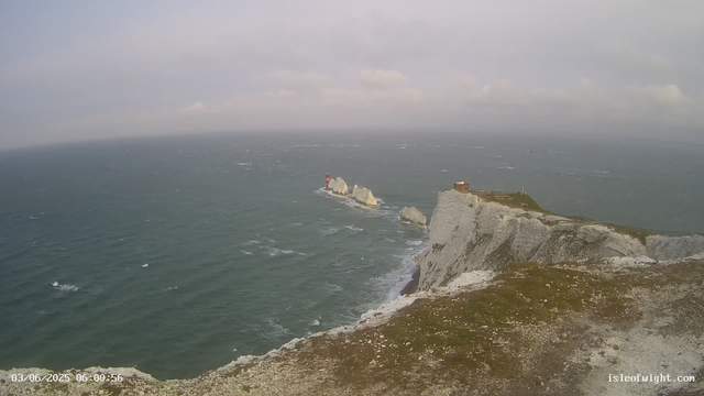 A view of a rugged coastline with white chalk cliffs extending into the sea. In the distance, there are smaller rock formations with a red and white lighthouse on one of them. The sky is mostly cloudy, and the ocean appears choppy with visible waves. There is a small building on top of the main cliff. The timestamp in the bottom left corner shows the date and time as "03/06/2025 06:00:56," and the URL "isleofwight.com" is displayed in the bottom right corner.