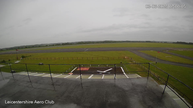 A view from a webcam at Leicester Aero Club shows an overcast sky with gray clouds. In the foreground, there is a helipad with a large white "H" on a red background and a white arrow pointing toward it. Surrounding the helipad is a black railing. Beyond the helipad, there is a grassy area, a small paved taxiway, and a runway stretching into the distance. There are some small signs and markings along the runway. The landscape is flat with trees visible on the horizon. The timestamp in the top right corner reads "03-06-2025 07:00:05". The bottom left corner displays the text "Leicestershire Aero Club".