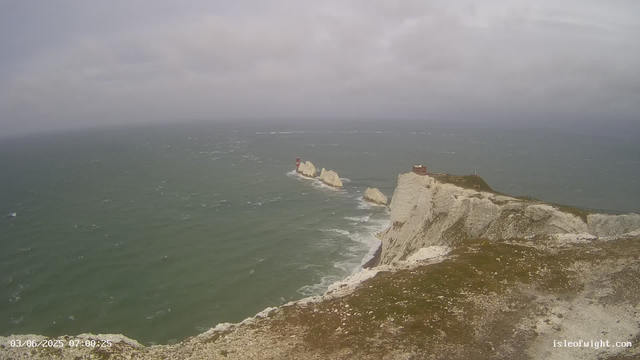A view of white chalk cliffs along the coast extending into the distance, with a rocky outcrop featuring a red and white lighthouse near the water. The sea appears choppy under a cloudy sky, and a small building is perched on the cliff edge. The photograph is timestamped "03/06/2025 07:00:25" and has the website "isleofwight.com" at the bottom right corner.