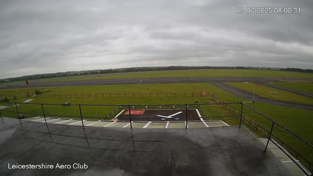 A view of an empty aircraft runway and surrounding grassy area, with overcast gray skies. In the foreground, a rooftop observation deck with a black railing and a helipad marked with a white helicopter symbol. The location is the Leicestershire Aero Club, as indicated by text at the bottom left corner. The scene appears to be captured early in the morning, with a timestamp showing "03-06-2025 08:00:31."