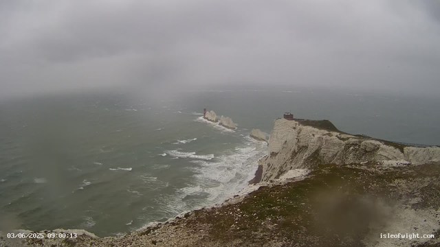 A view of white chalk cliffs with a lighthouse and a small building on top, overlooking the choppy sea under a cloudy, overcast sky. Waves are breaking against the rocks below, and the landscape is shrouded in mist, giving a moody, stormy atmosphere. The timestamp on the bottom left reads "03/06/2025 09:00:13," and the website "isleofwight.com" is shown on the bottom right.