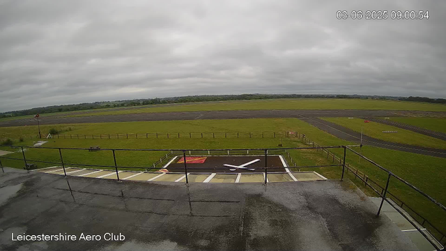 An outdoor view from a webcam at Leicester Aero Club shows a cloudy sky over a runway and grassy area. In the foreground, there is a fence, a small helipad with a white H and a red square, and a few small objects on the ground. The background consists of an open grassy and paved area with no aircraft visible, and a distant tree line. The timestamp indicates the date and time as June 3, 2025, at 9:00 AM.