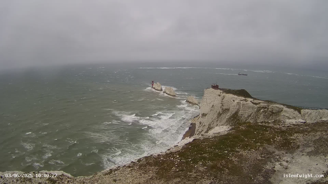A view of rugged white chalk cliffs overlooking the sea on a cloudy day. The ocean is choppy with small waves, and there are several large rocks and a small lighthouse on the rocks near the shoreline. A boat is visible in the distance, leaving a wake behind it. The landscape appears wild and windswept.
