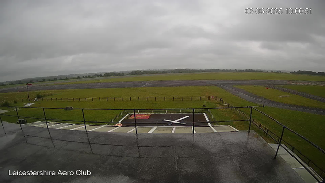 An overcast view of a small airfield under gray, cloudy skies. In the foreground, there is a helicopter landing pad with a white "X" marking and a red background. The airfield has several runways and taxiways extending into the distance. A windsock is visible on the left side, and there's a grassy area enclosed by a wooden fence. The landscape beyond the airfield is flat with fields and trees on the horizon. The timestamp in the upper right corner reads "03-06-2025 10:00:51" and the bottom left corner says "Leicestershire Aero Club."