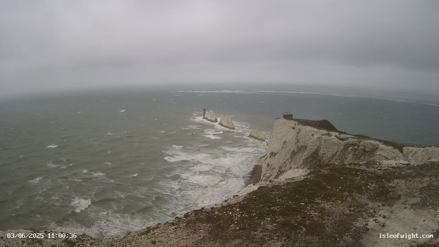 A view of a rugged coastline with steep white chalk cliffs extending into the ocean. The sea appears choppy with visible waves. On top of the cliffs, there is a small building or shelter near the edge. In the distance, there are rock formations protruding from the water, with one of them bearing a red and white lighthouse. The sky is overcast, giving a gray and gloomy atmosphere. The image has a date and time stamp at the bottom left corner and a website URL at the bottom right corner.