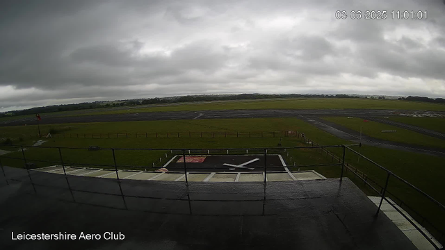 A view of the Leicestershrie Aero Club airfield under a cloudy sky, with a small helicopter landing pad in the foreground and the runway stretching into the distance. The landscape is flat and green, with some remote trees and fields beyond the airfield. The timestamp indicates the date as June 3, 2025, at 11:01 AM.