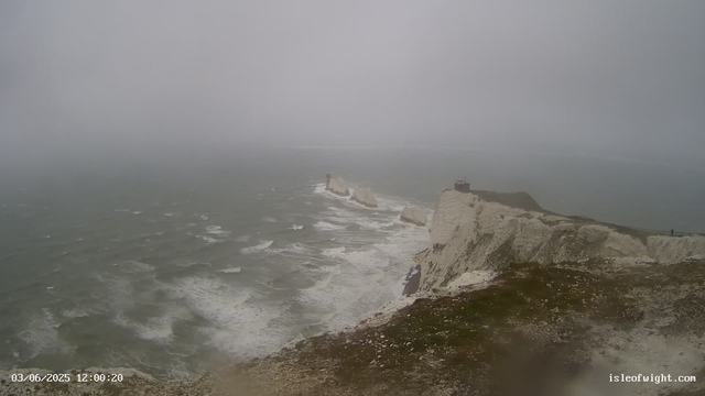 A view of the white chalk cliffs at Étretat on a foggy day, with the cliffs extending into the ocean. The sea appears choppy with waves crashing against the base of the cliffs, and the sky is overcast with thick fog obscuring the distance. There are small figures of people visible in the distance near the top of the cliffs. The image has a timestamp of "03/06/2025 12:00:20" and the website "isleofwight.com" at the bottom right corner.