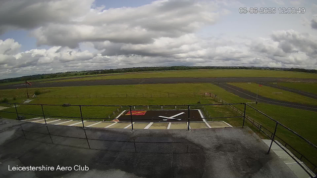 A wide view of an airport tarmac and surrounding grassy fields under a partly cloudy sky. The photo includes a viewing platform with a railing in the foreground and a marked helicopter landing pad. The scene is calm with no aircraft visible on the runway. The timestamp indicates the photo was taken on June 3, 2025, at 12:00 PM.