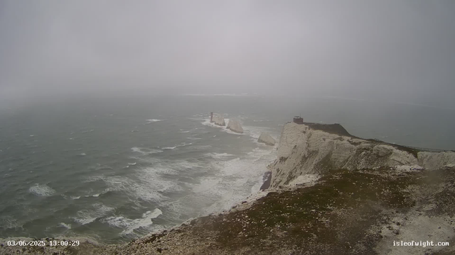 A foggy view of white chalk cliffs with a lighthouse on top, extending into the ocean with waves crashing against the base of the cliffs. There are some rocks visible offshore in the water. The scene appears to be on a cloudy, misty day, with limited visibility. The image includes a time and date stamp in the lower left corner and a website URL in the lower right corner.