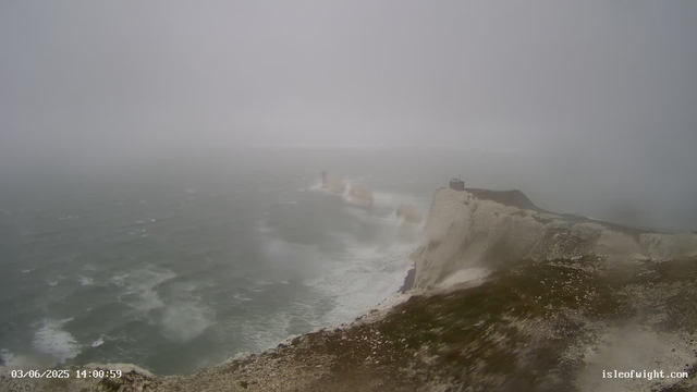 A foggy seascape showing white chalk cliffs with a small building on top, overlooking turbulent waters below. Waves crash against the base of the cliffs in a misty, overcast atmosphere.