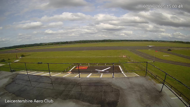 A view from a webcam at Leicestershire Aero Club shows a grassy airfield with runways extending into the distance under a partly cloudy sky. In the foreground, there's a section with a large letter "X" painted on the ground, surrounded by a railing, and a red and yellow marked area. The scene includes a windsock on the left, a small vehicle on the runway, and a distant airplane on the tarmac. The timestamp indicates the date and time as June 3, 2025, at 15:00:48.