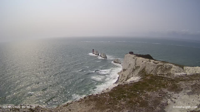 A view of white chalk cliffs extending into the ocean with a small building on top of the cliff. In the distance, rock formations and sea stacks are visible emerging from the water. The sky is overcast with a hazy atmosphere. The image is timestamped "03/06/2025 16:00:28" in the lower left corner.