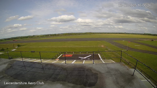 A view from a webcam at Leicesterhire Aero Club showing a runway and surrounding grassy area under a partly cloudy sky. In the foreground, there is a platform with a fenced edge and a helicopter landing pad marked with a large white "X" and a red and yellow square. The horizon shows trees and open fields in the distance. The date and time displayed in the top right corner are "03-06-2025 16:00:26".