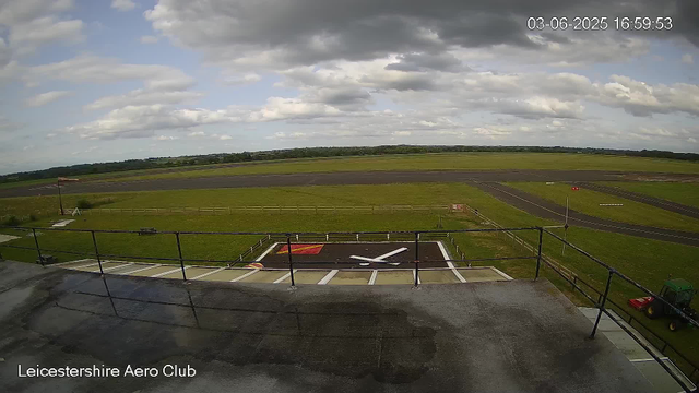 A view of an airfield under partly cloudy skies, with runways stretching into the distance. In the foreground, a helipad marked with a large white “H” on a red background is visible, surrounded by a white railing and some equipment. A small tractor is parked along the edge of the helipad. Text at the bottom left reads “Leicestershire Aero Club”. The image is timestamped “03-06-2025 16:59:53” in the top right corner.