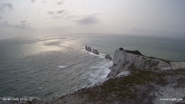 A view of white chalk cliffs along the coastline extending into the ocean, with the sea surrounding the base of the cliffs. The sky is overcast with gray clouds, and the sunlight reflects off the water's surface. There is a structure on the edge of the cliff and a path along the top. The image is timestamped "03/06/2025 17:01:12" and has the website "isleofwight.com" in the bottom right corner.