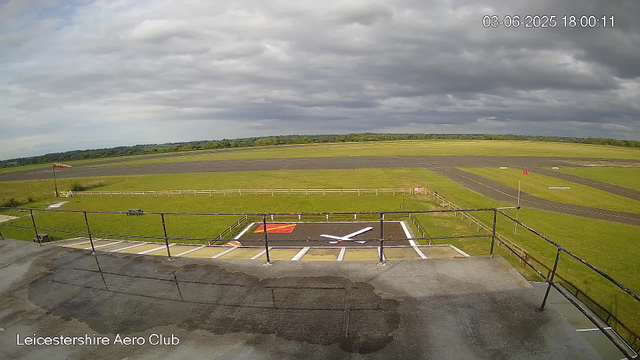 A view from a webcam at Leicesterhire Aero Club shows an empty aircraft parking area with a marked helipad in the foreground. Beyond the parking area, a grassy field surrounds a runway and taxiways, extending towards a horizon under a cloudy sky. The image is timestamped June 3, 2025, at 18:00:11. A windsock is visible on the left side, indicating wind direction, and there are a few small barriers and objects scattered around. The overall scene is calm and overcast.