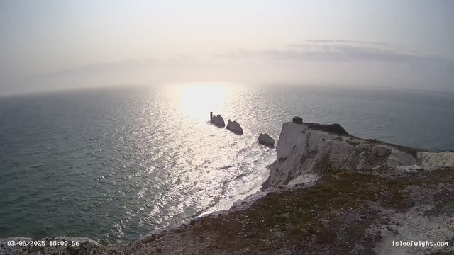 A view of white chalk cliffs overlooking the ocean during sunset, with the sun low on the horizon casting a bright reflection on the water. The cliffs are rugged with some dark structures at the top, and several large sea stacks stand offshore in the distance. The sky is partly cloudy, and the overall scene is serene and picturesque.