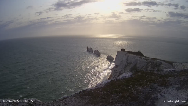 A photograph taken from a webcam shows a coastal scene during sunset or sunrise. The sky is partly cloudy with the sun low on the horizon, casting a golden light on the water and cliffs. The ocean appears calm with gentle waves, and there are white chalk cliffs with dark patches along the shoreline. Several large rock formations protrude from the water near the base of the cliffs. At the very top of the cliffs, there is a small structure, possibly a lighthouse or a small building, with a pathway leading to it. The sky has a mix of clouds, and the overall atmosphere is serene and picturesque. There is a timestamp on the bottom left corner indicating the date as March 6, 2025, at 19:00:05, and a website URL in the bottom right corner: isleofwight.com.
