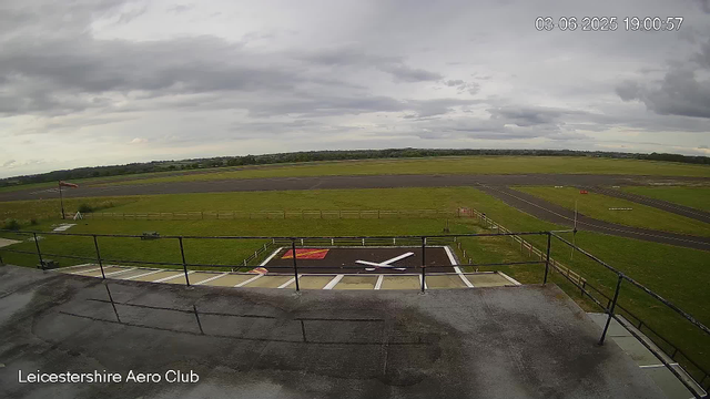 An overcast sky with dark, cloudy weather at Leicesterhire Aero Club. The image shows the runway and grassy areas surrounding it, with a windsock on the left and a small helicopter landing pad in the foreground. The scene appears calm with no aircraft visible on the ground.