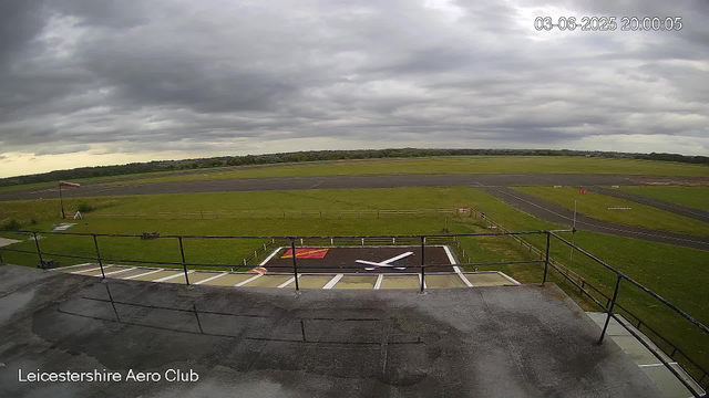 A view from a webcam at Leicestershire Aero Club showing an overcast sky with dark gray clouds. In the foreground, there is a rooftop with a railing and a helicopter landing pad marked with a red square and a white "H." The open grassy area surrounds the runway, which extends into the distance. There is a flagpole with a flag to the left and some small structures and signs near the runway. The scene is calm with no aircraft or people visible. The timestamp indicates the image was captured on June 3, 2025, at 8:00 pm.