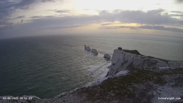 A view of the white chalk cliffs along the coastline at sunset, with the sea stretching out below. There are several large rock formations in the water near the cliffs. A lighthouse stands atop the cliff on the right side of the image. The sky is partly cloudy, with the sun setting in the horizon. The image is timestamped "03/06/2025 20:01:09" and includes the URL "isleofwight.com".