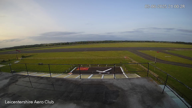 An outdoor view from a webcam at Leicesterhire Aero Club. The image shows a runway with a runway clock in the foreground, the sky is partly cloudy, and the landscape extends to the horizon with fields and trees visible in the distance. The timestamp in the top right corner indicates the date and time as June 3, 2025, at 21:00:28.