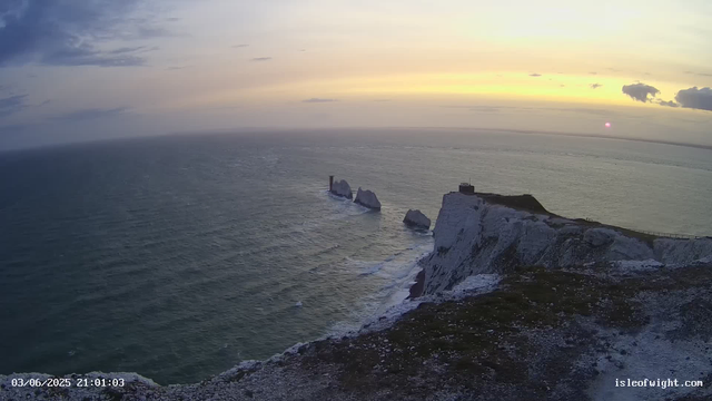 A scenic view of white chalk cliffs overlooking the ocean during sunset, with the sun near the horizon casting a warm glow. The cliffs have grassy tops, and there are large rock formations in the water nearby. The sky is partly cloudy with a mix of light and darker clouds. The image includes a timestamp at the bottom left corner, reading "03/06/2025 21:01:03," and the text "isleofwight.com" at the bottom right corner.