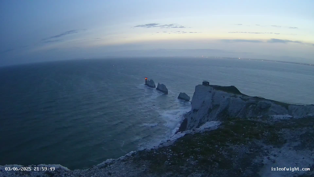 A view of white chalk cliffs overlooking the sea during dusk. A lighthouse with a red light is situated on smaller rock formations jutting out from the water, and there is a building on the largest cliff. The sky is partly cloudy with the horizon line visible in the distance. The timestamp in the bottom left corner reads "03/06/2025 21:59:49," and the website "isleofwight.com" is displayed in the bottom right corner.