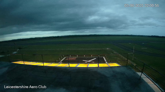 A wide view of an airfield or aerodrome at dusk or dawn, with a large helicopter landing pad marked by a white circle and a prominent letter "H" in the center. The sky is cloudy and dark, suggesting overcast weather. In the distance, there are open fields or grasslands stretching to the horizon. A platform or observation deck with a black railing is in the foreground, and some illuminated yellow sections can be seen on the platform. The image includes a timestamp in the upper right corner and text reading "Leicestershire Aero Club" in the lower left corner.