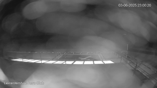 A black-and-white, nighttime view of Leicester Aero Club taken from a webcam. The image shows a large, open space with a runway or tarmac area, and a runway light or signal towards the center bottom. There is a fence or railing near the camera. The sky appears cloudy with some light patches. The timestamp on the image indicates it was taken on June 3, 2025, at 11:00 PM.