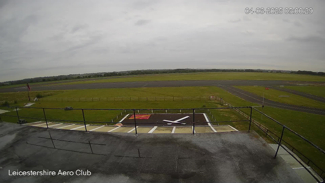 An empty outdoor airfield with a paved runway and grassy areas. There is a small helipad marked with a red and white symbol, and a windsock flying on a pole near the left side. A balcony with a black railing is visible in the foreground. The sky is overcast with gray clouds. The timestamp shows the date as June 4, 2025, at 5:00 AM. The location is the Leicestershire Aero Club.