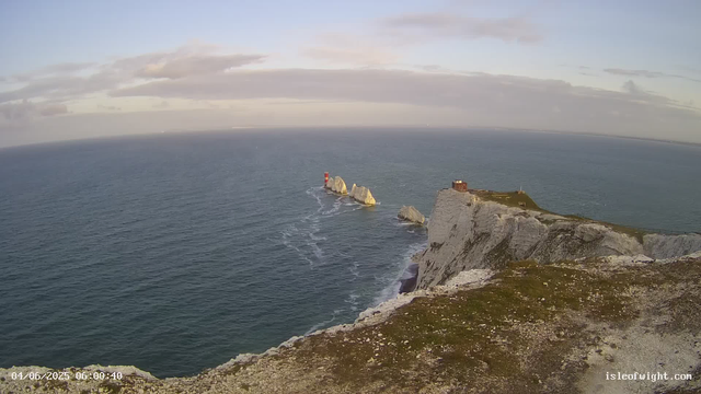 An aerial view of a rugged coastal landscape showing tall white chalk cliffs with patches of green grass on top. In the ocean below, large rocks and a small lighthouse with red and white stripes are visible near the shoreline. The sky is partly cloudy with a mix of soft light and shadows. The scene appears to be early morning or late afternoon. The image shows the date and time as 04/06/2025 at 06:00:40 and a website URL "isleofwight.com" in the bottom right corner.