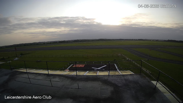 An outdoor view from a webcam at Leicestershire Aero Club shows a cloudy sky with the sun low on the horizon. In the foreground, there is a helipad with a large white "H" symbol, surrounded by a safety fence. Beyond the helipad, there are grassy fields and a paved airstrip with markings, along with some small structures and runways in the distance. The timestamp in the top right corner indicates the date and time as April 6, 2025, at 6:00:59 AM.