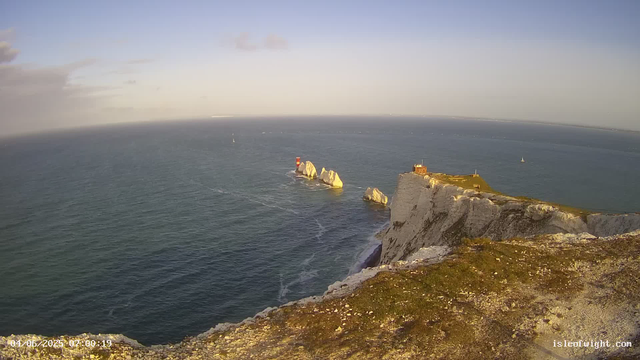 A view of a rugged coastline with white chalk cliffs topped with green grass. In the water near the cliffs, there are several large rock formations, some with a lighthouse on top. The sky is mostly clear with a few clouds, and a few sailboats are visible in the distance. The photo was taken in the early morning.