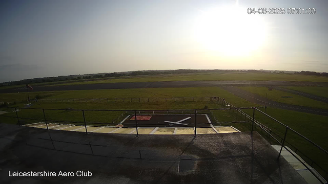 An outdoor view from a surveillance camera at Leicesterhire Aero Club showing a runway and surrounding grassy area. The sky is clear with the sun shining brightly, suggesting early morning. There is a black helicopter hangar or shelter with a helipad in the foreground. A windsock is visible on the left side near the fence. The timestamp in the upper right corner indicates the date as April 6, 2025, and the time as 07:01.