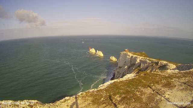 A scenic aerial view of white chalk cliffs along the coastline, with a grassy top and a small building or lighthouse at the edge. The cliffs overlook the ocean, which has several large rock formations and a red and white striped lighthouse on one of the rocks in the water. A sailboat is visible on the horizon, and the sky is partly cloudy with sunlight illuminating the scene.