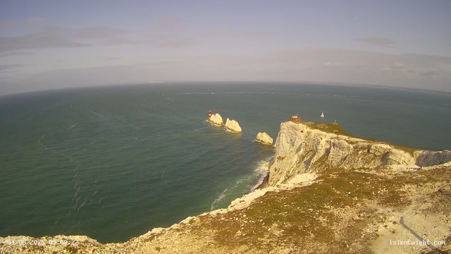 Aerial view of white chalk cliffs extending into the ocean with a lighthouse on a cliff top, a small building nearby, and sailboats in the distance. The sea has visible wave patterns and a sailboat is sailing close to the cliffs. The sky is partly cloudy with soft sunlight illuminating the scene.
