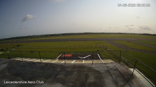 The image shows an outdoor view from a webcam at Leicestershire Aero Club, taken on June 4, 2025, at 9:00 in the morning. In the foreground, there is a rooftop with a wind sock, some railings, and a helipad marked with a large "H." Beyond the rooftop, there is a grassy area enclosed by a wooden fence, with a windsock on a pole to the left. Further back, there are runways and taxiways, with a vast open field in the distance under a partly cloudy sky.