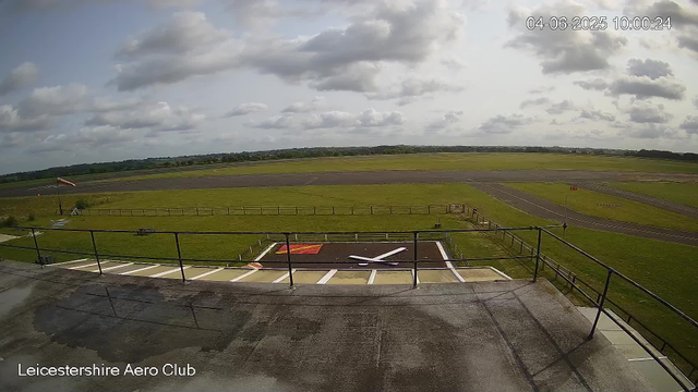 An overcast day at Leicestershire Aero Club visible from a webcam. The foreground shows a small helicopter pad with a white helicopter silhouette painted on the ground. There are parking spaces with white lines, and a railing surrounds the area. In the background, there are open grassy fields, taxiways, and runways extending into the distance. The sky is filled with scattered clouds, and there is a windsock on the left side of the image. The timestamp reads 04-06-2025 10:00:24.