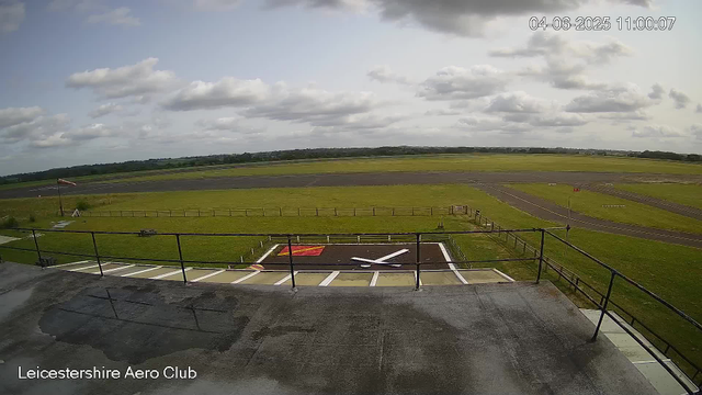 A view from a webcam at Leicesterhire Aero Club showing an outdoor space with a small tarmac area in the foreground, enclosed by a black metal railing. There is a painted landing or parking zone marked with white and red lines. Beyond this area, the scene extends to a grassy field with a windsock on a pole and a windsock flag, indicating outdoor conditions. The background features an expansive, flat landscape with an open sky filled with scattered clouds. The timestamp on the image reads "04-06-2025 11:00:07".