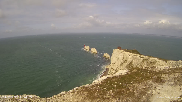 A view of white chalk cliffs overlooking the ocean. Several large rocks are situated in the water near the base of the cliffs, with a red and white lighthouse on one of the rocks. A small building is perched on the edge of the cliff to the right. The sky is partly cloudy with patches of sunlight.