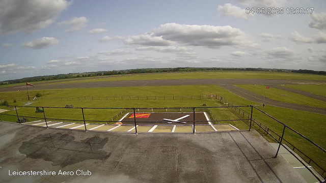 An outdoor view from a webcam at Leicestershire Aero Club shows a section of a tarmac area with painted markings and a red and white windsock on a pole to the left. Beyond the tarmac, a grassy field and a runway extend towards the horizon under a partly cloudy sky. There are some small structures and equipment on the grass, and a fence surrounds the area. The scene is calm with no visible aircraft or people. The timestamp indicates the date is April 6, 2025, and the time is 12:00:17.