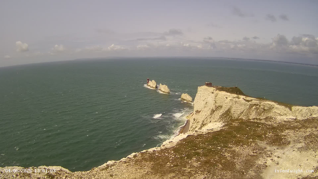 Aerial view of white chalk cliffs with a small building on top, extending into the sea. In the water, several large rock formations and a red and white lighthouse on one of the rocks are visible. The sky is partly cloudy with patches of blue.